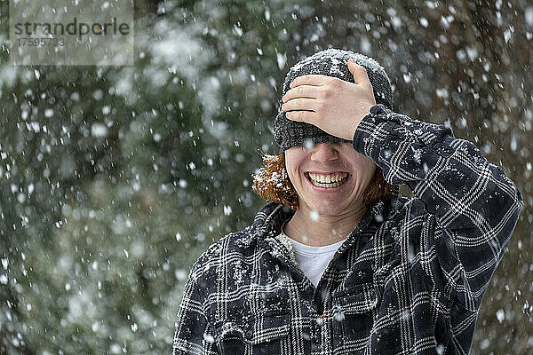 Fröhlicher Mann bedeckt Augen mit Strickmütze im Schneefall