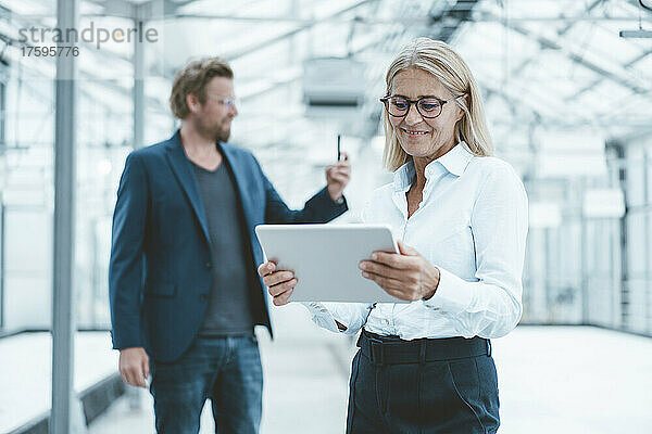 Lächelnde Geschäftsfrau nutzt Tablet-PC mit Kollege im Hintergrund im Büro