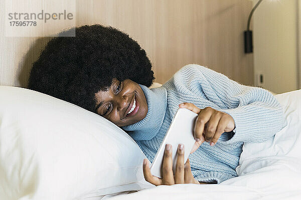 Lächelnde Frau mit Rollkragenpullover  die zu Hause im Schlafzimmer auf dem Smartphone zuschaut