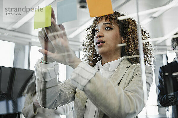Junge Geschäftsfrau mit braunem lockigem Haar klebt im Büro Klebezettel an die Glaswand