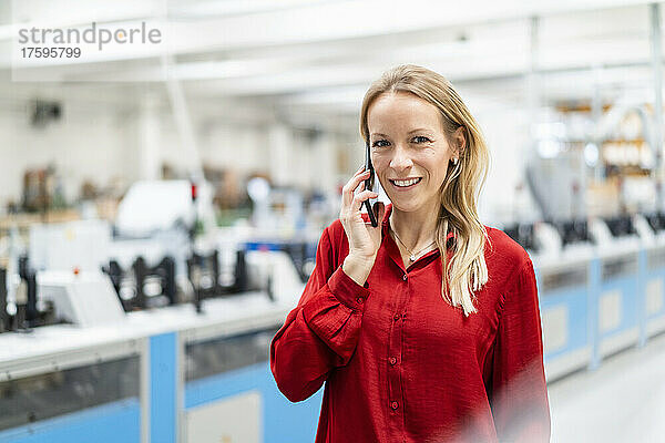 Lächelnde blonde Geschäftsfrau  die in der Fabrik mit dem Smartphone spricht