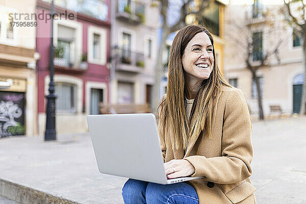 Glückliche Frau mit Laptop sitzt auf Fußweg in der Stadt