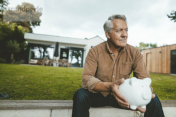 Nachdenklich lächelnder älterer Mann mit Sparschwein sitzt im Hinterhof