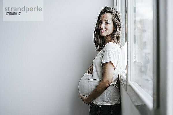 Lächelnde schwangere Frau lehnt zu Hause am Fenster
