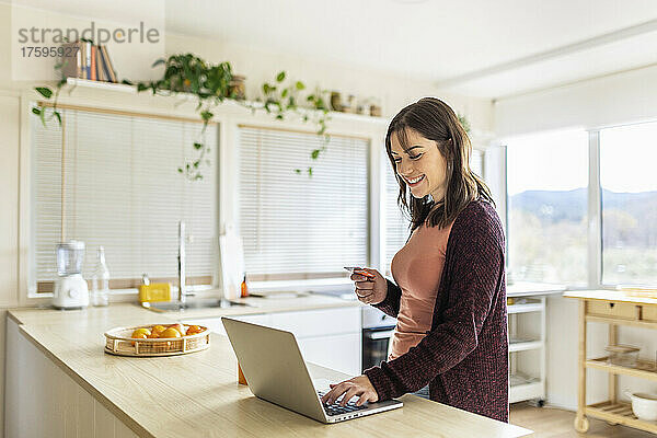 Geschäftsfrau macht Online-Einkäufe am Laptop und hält Kreditkarte in der heimischen Küche