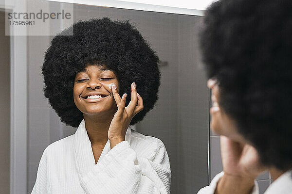 Lächelnde Frau trägt im Badezimmer Gesichtscreme auf die Wange auf