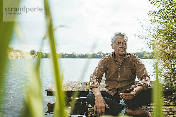 Nachdenklicher älterer Mann mit Mobiltelefon sitzt auf einem Steg am See