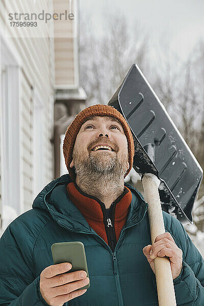 Glücklicher Mann mit Smartphone und Schneeschaufel  der im Winter nach oben schaut