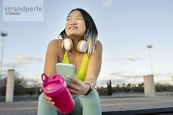 Smiling woman with smart phone and water bottle on bench