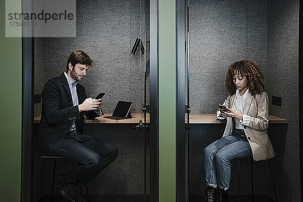 Geschäftsmann und Geschäftsfrau sitzen am Schreibtisch in der Kabine und nutzen Smartphones
