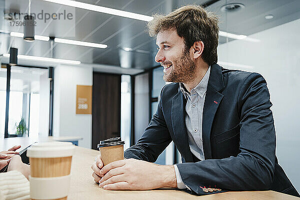 Smiling businessman holding disposable coffee cup enjoying coffee break in office