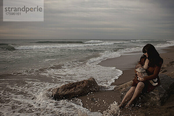 Mother breastfeeding newborn baby boy sitting on rock at beach