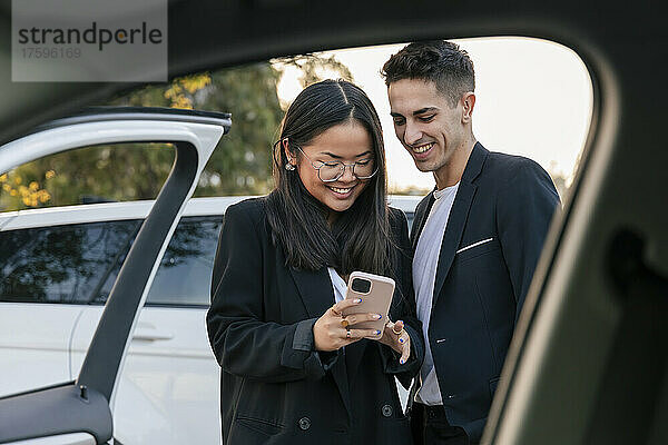 Lächelnde Geschäftskollegen teilen ihr Smartphone vor dem Auto