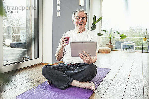 Lächelnder älterer Mann sitzt mit einem Glas Saft und einem Tablet-PC zu Hause auf der Trainingsmatte