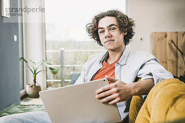 Lächelnder Mann mit Laptop sitzt auf dem Sofa im Wohnzimmer