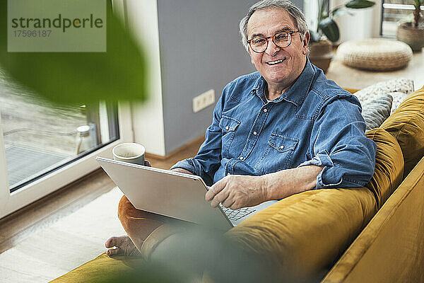 Lächelnder älterer Mann mit Laptop und Kaffeetasse sitzt zu Hause auf dem Sofa