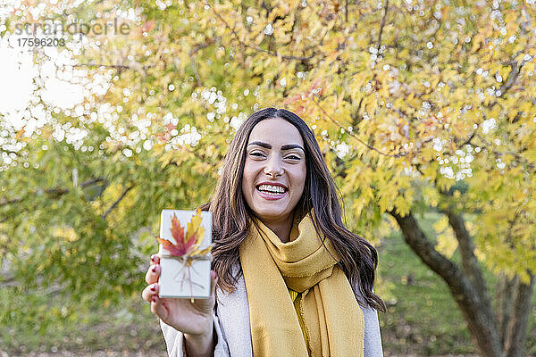 Fröhliche junge Frau hält kleine Geschenkbox mit Herbstblatt im Park