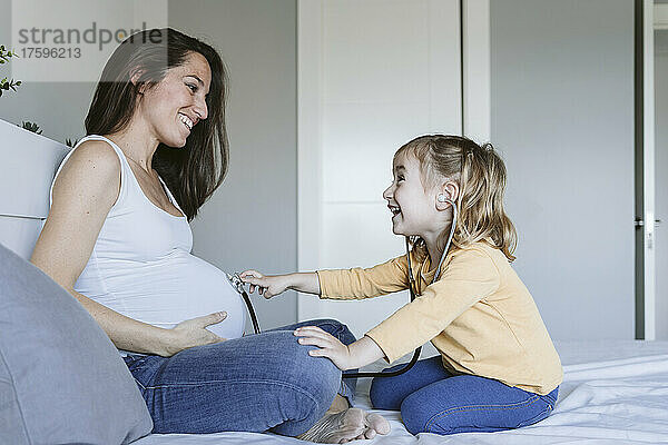 Glückliches Mädchen  das durch Stethoskop auf dem Bett dem Bauch der schwangeren Mutter zuhört