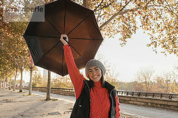 Lächelnde junge Frau mit Regenschirm steht auf Fußweg