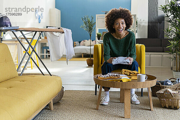 Glückliche Afro-Frau faltet Kleidung  die zu Hause auf einem Stuhl im Wohnzimmer sitzt
