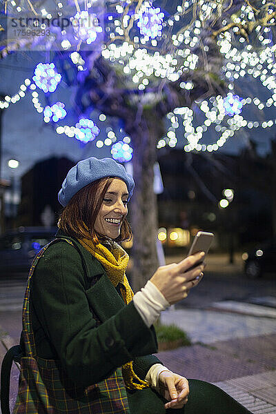 Glückliche Frau  die nachts vor einem beleuchteten Baum sitzt und Textnachrichten per Smartphone sendet