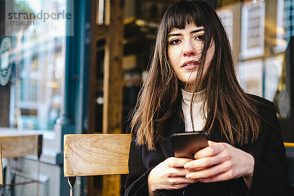 Schöne junge Frau mit Pony hält Smartphone im Straßencafé in der Hand