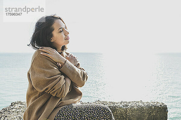 Junge Frau umarmt sich selbst und sitzt auf einem Felsen am Strand