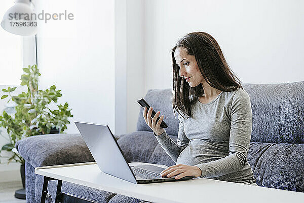Schwangere Frau mit Smartphone arbeitet im Heimbüro am Laptop