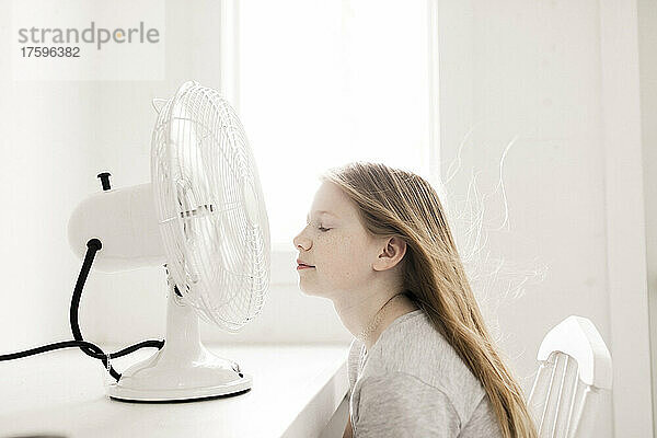 Mädchen sitzt zu Hause vor einem Tisch mit elektrischem Ventilator