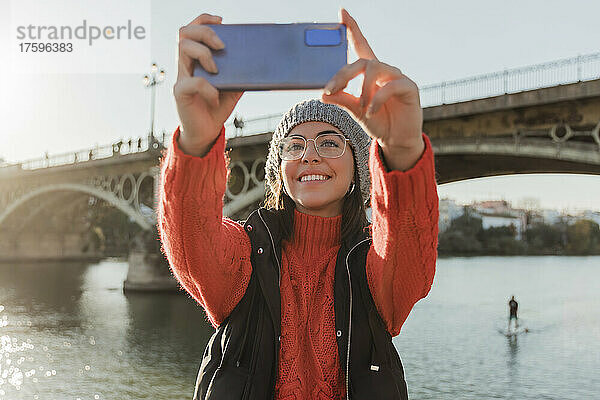 Happy beautiful woman taking selfie with mobile phone in front of bridge