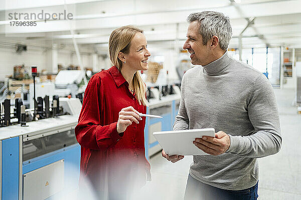 Glückliche blonde Geschäftsfrau tauscht Ideen mit Kollegen aus  die in der Fabrik einen Tablet-PC halten