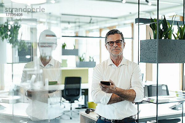 Lächelnder Geschäftsmann mit Smartphone  der neben dem Regal im Büro steht
