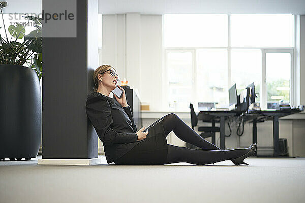 Geschäftsfrau spricht mit Smartphone und sitzt im Büro auf dem Boden
