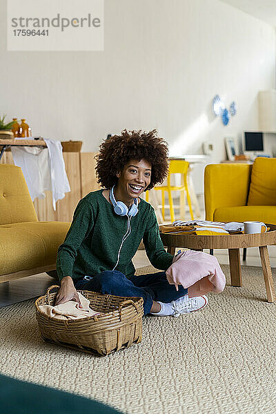 Glückliche Frau mit lockigem Haar  die Kleidung faltet  sitzt auf dem Teppich im Wohnzimmer