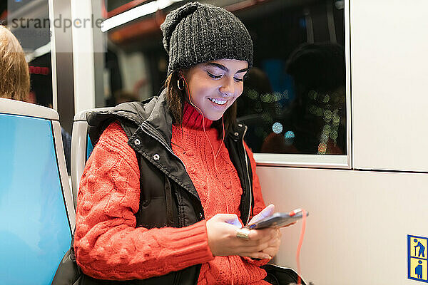 Lächelnde junge Frau benutzt Smartphone und hört Musik über In-Ear-Kopfhörer in der Straßenbahn