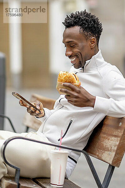 Lächelnder Mann mit Burger und Mobiltelefon auf Bank sitzend