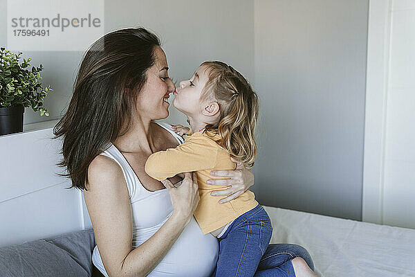 Süßes Mädchen küsst die Nase der schwangeren Mutter zu Hause im Bett