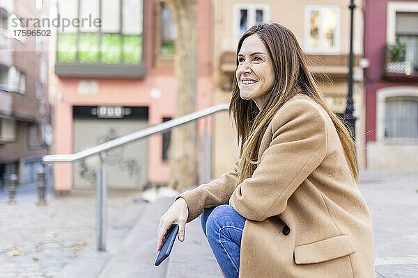 Lächelnde Frau mit Smartphone sitzt auf Fußweg in der Stadt