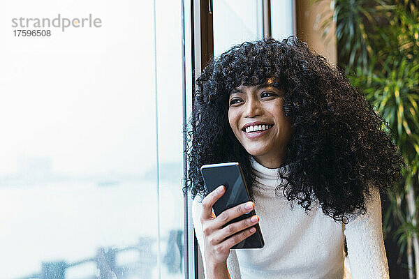 Nachdenkliche junge Frau hält lächelnd ihr Mobiltelefon am Fenster eines Cafés