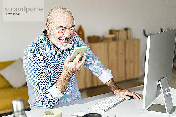 Lächelnder älterer Geschäftsmann  der am Schreibtisch Voicemail über sein Smartphone sendet