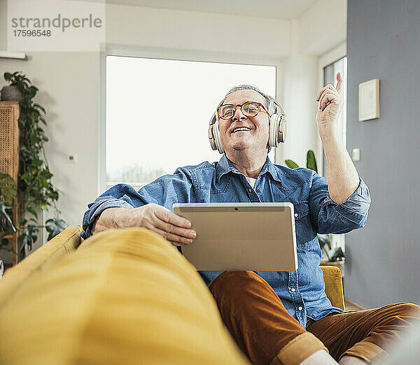 Glücklicher älterer Mann hält Tablet-PC in der Hand und genießt Musik über kabellose Kopfhörer im Wohnzimmer