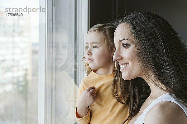 Lächelnde Frau mit Tochter  die zu Hause durch das Fenster schaut