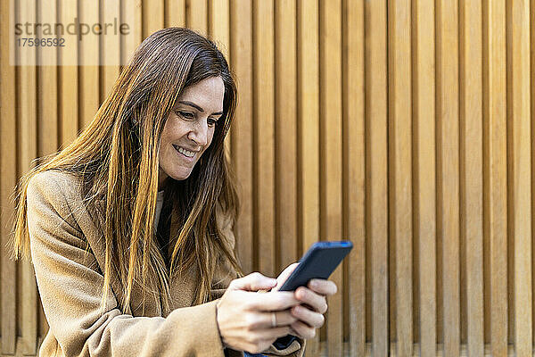 Lächelnde Frau  die Textnachrichten über ihr Smartphone vor der Wand sendet