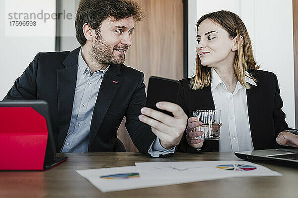Geschäftsfrau schaut Kollegen mit Smartphone an  die am Schreibtisch im Büro diskutieren