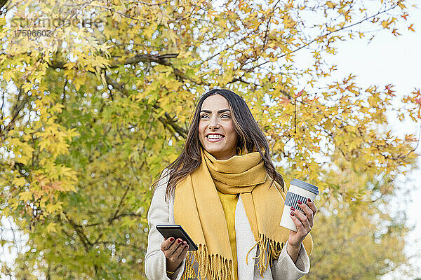Lächelnde junge Frau mit Smartphone und wiederverwendbarer Kaffeetasse im Herbstpark