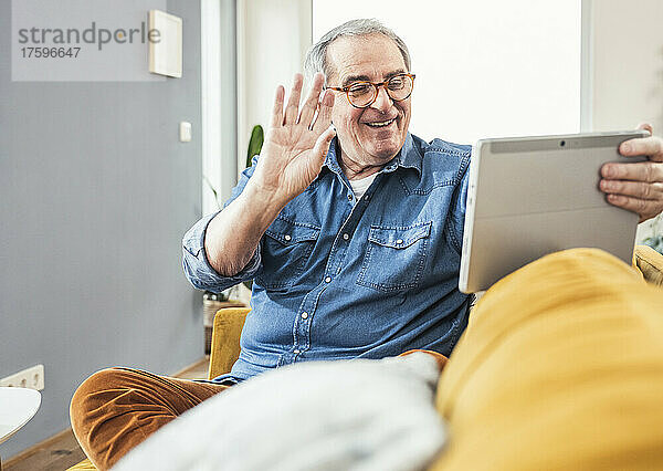 Lächelnder Mann winkt bei einem Videoanruf über einen Tablet-PC im heimischen Wohnzimmer