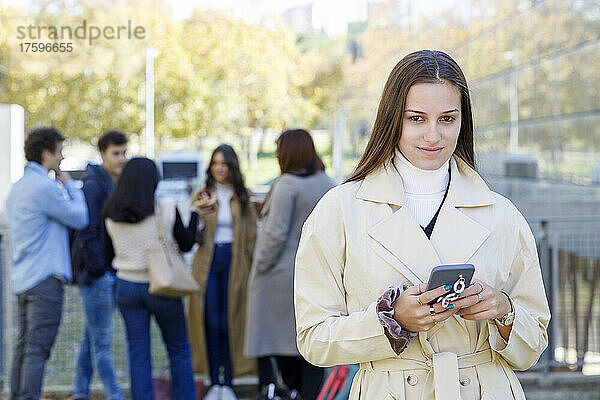 Junge Frau hält Mobiltelefon mit Freunden im Hintergrund auf dem College-Campus