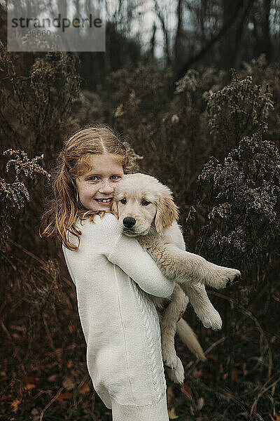 Lächelndes Mädchen mit Golden Retriever-Welpe steht vor Baum