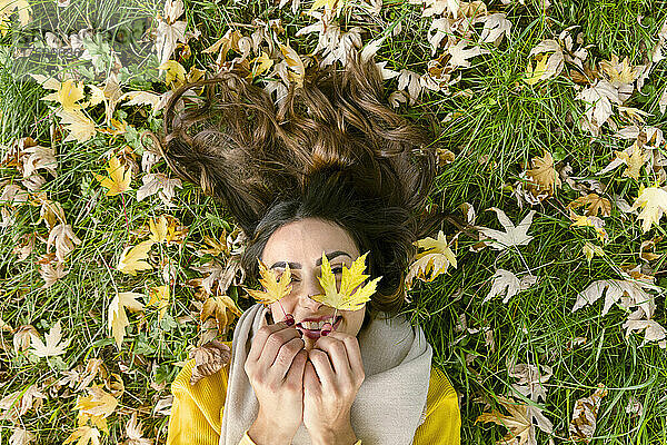 Happy young woman lying on grass holding autumn leaves in park