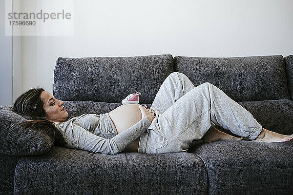 Schwangere Frau mit Babyschuhen auf dem Bauch liegt zu Hause auf dem Sofa